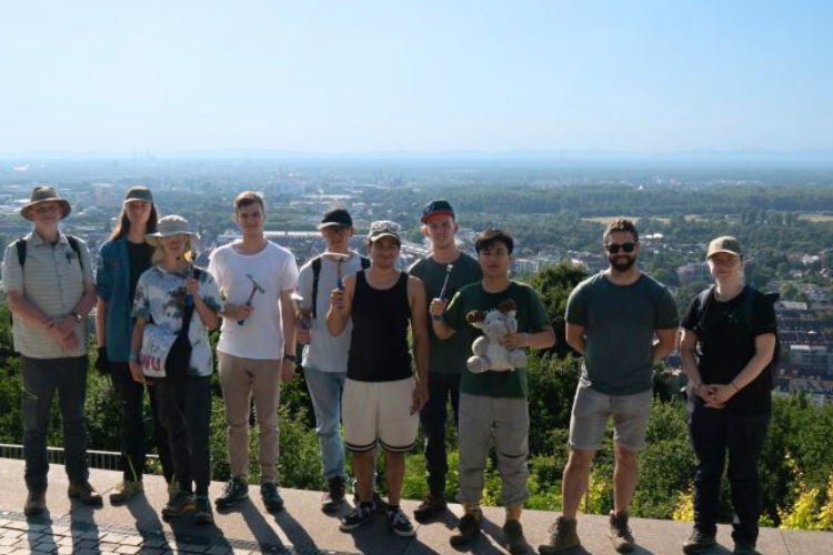 Gruppe junger Menschen mit geologischen Hämmern vor einer Stadtpanorama-Aussicht.