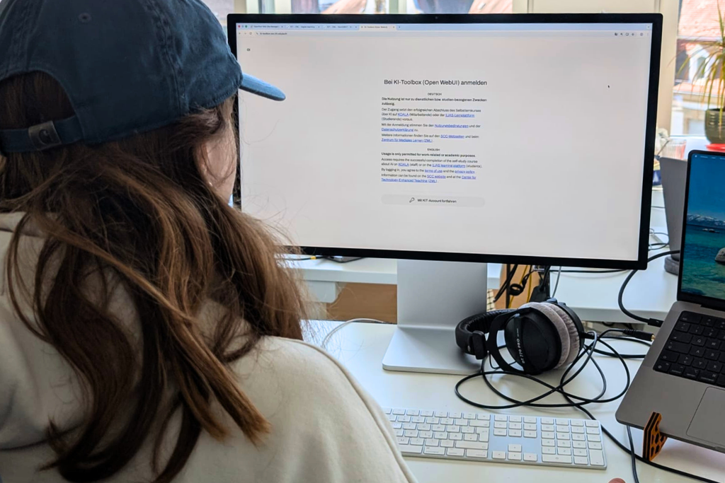 Person sitzt am Schreibtisch vor einem großen Monitor, Kopfhörer, Tastatur und MacBook.