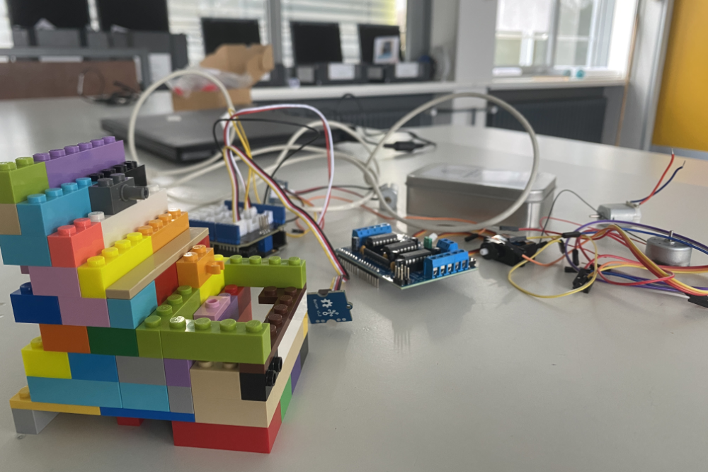 Bunte LEGO-Steine zu einem mehrfarbigen Blockturm auf einem Labor-/Arbeitsbench mit Elektronik-Boards und Kabeln.